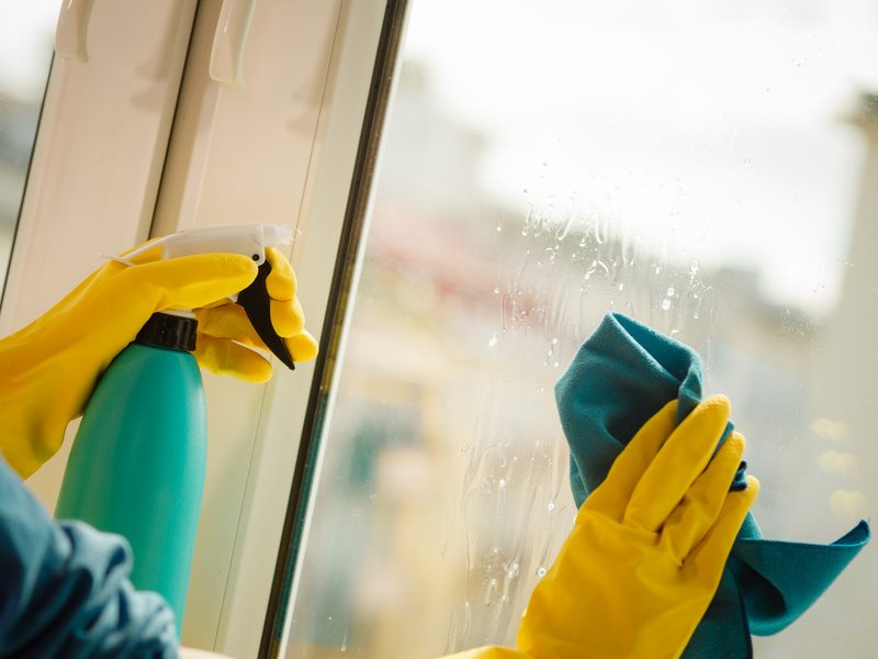 Female hand in yellow gloves cleaning window with blue rag and spray detergent. Spring cleanup, housework concept